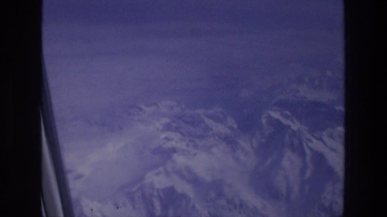 Snowy mountain peaks seen through a dark airplane window frame under a purple sky.