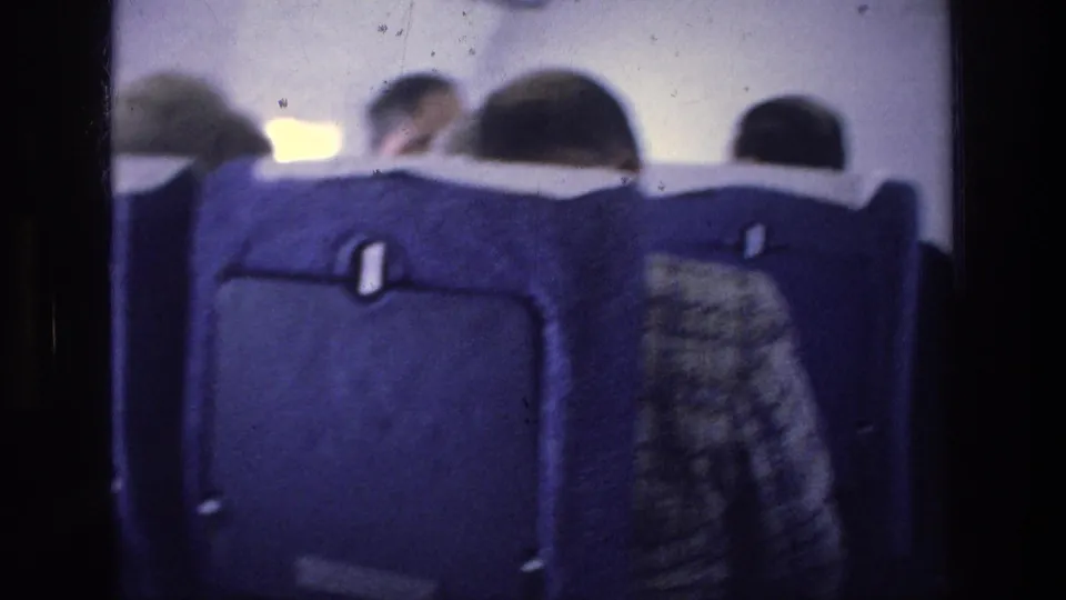 Inside the aircraft cabin, blue seat backs and several seated passengers appear in soft 1970s 8mm home-movie footage.