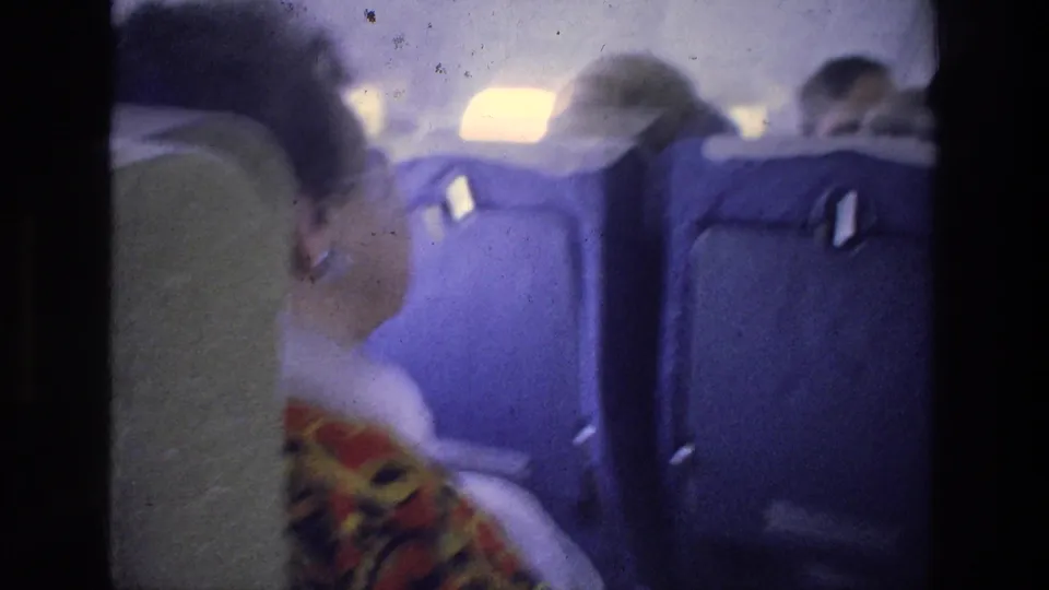 A handheld 8mm view looks across nearby passengers and blue aircraft seats during the 1976 Athens travel flight.
