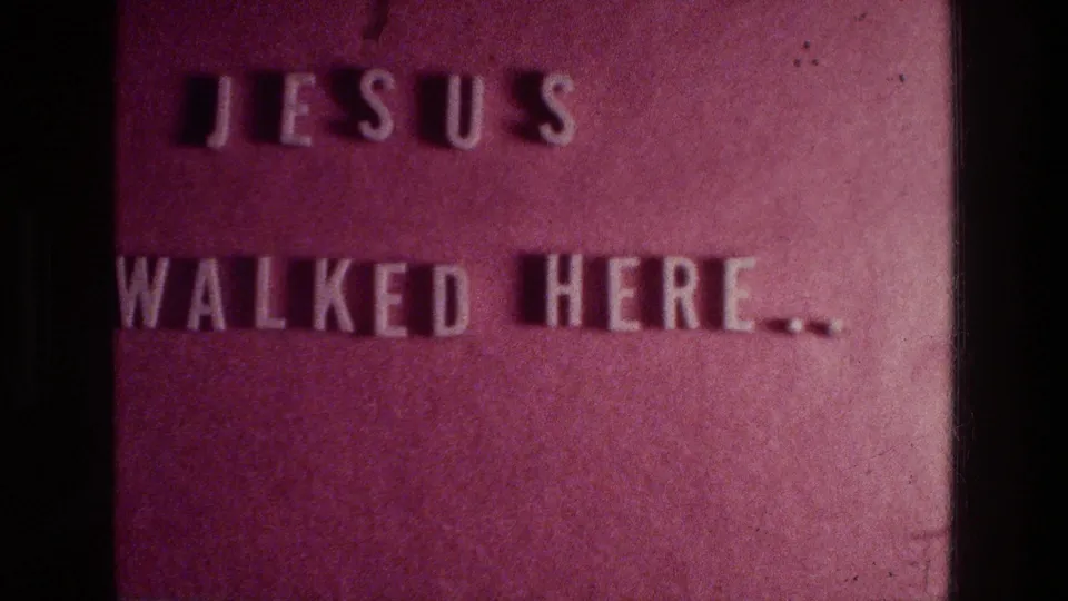 A 1976 Athens home-movie frame holds on the reddish embossed plaque text, with dark borders and visible 8mm grain.