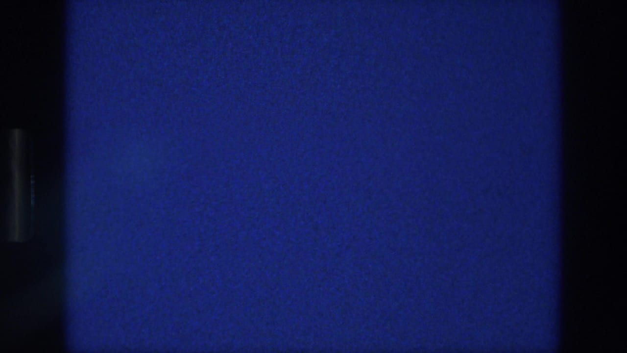 1976 Athens 8mm frame with a static blue field, fine analog grain, light surface specks, and a dark vertical divider along the left side.