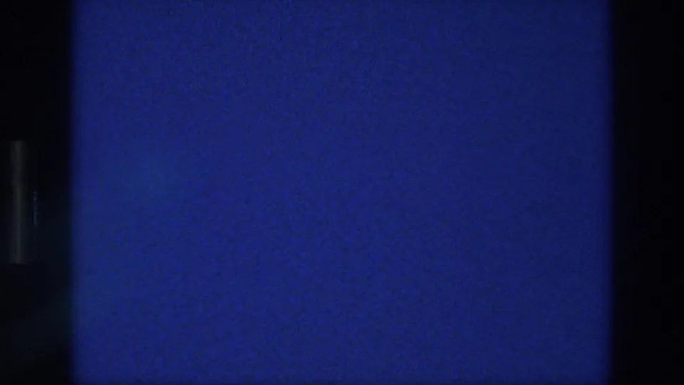1976 Athens 8mm frame with a static blue field, fine analog grain, light surface specks, and a dark vertical divider along the left side.