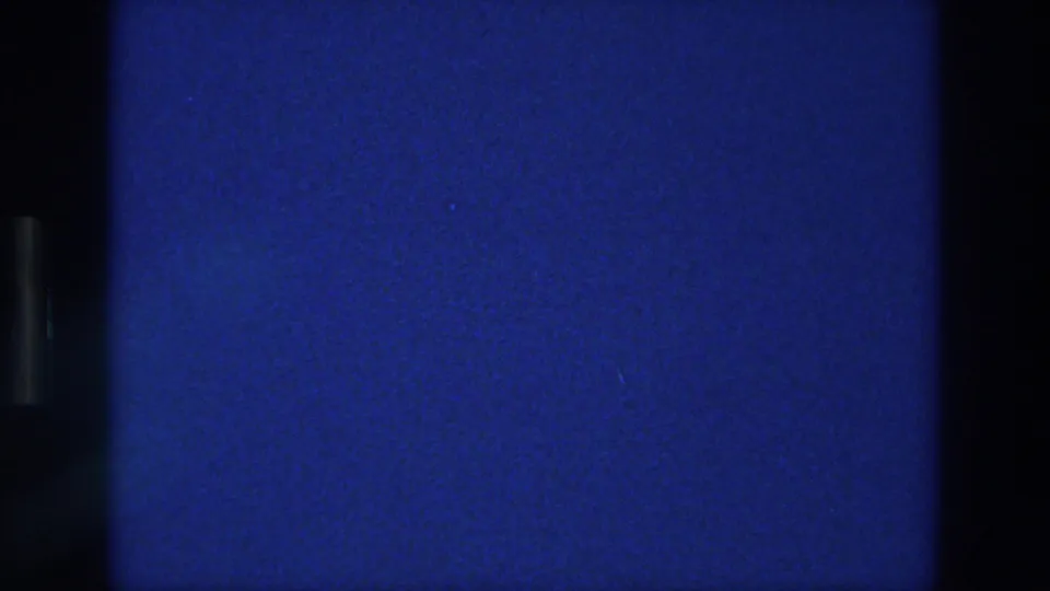 1976 Athens 8mm frame showing a blank saturated blue area with film grain, dust flecks, dark borders, and slight tonal variation.
