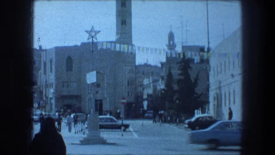 1976 8mm Bethlehem street scene with pedestrians, passing cars, a tower, hanging banners, and a large star symbol on a building.