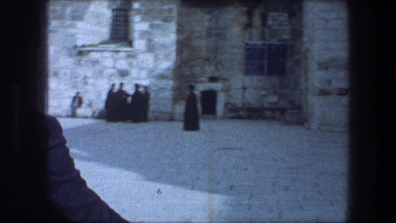 1976 8mm frame from Bethlehem showing a stone courtyard, dark-robed figures near a building, and a cropped visitor in the foreground.