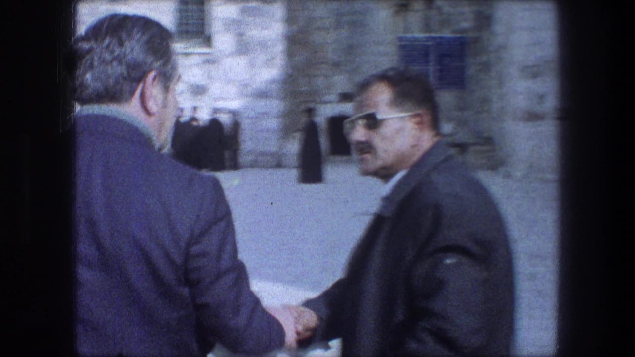 Two men in suits shake hands in a stone courtyard, with robed figures in the background.