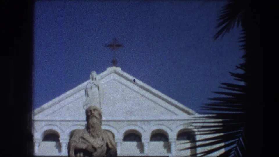 1976 8mm view in Bethlehem of a white church facade with cross, statue, arched details, and palm fronds at the edge.
