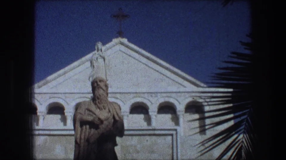 Bethlehem 1976 home-movie frame showing a church facade topped by a cross, with a central statue and palm fronds along the side.