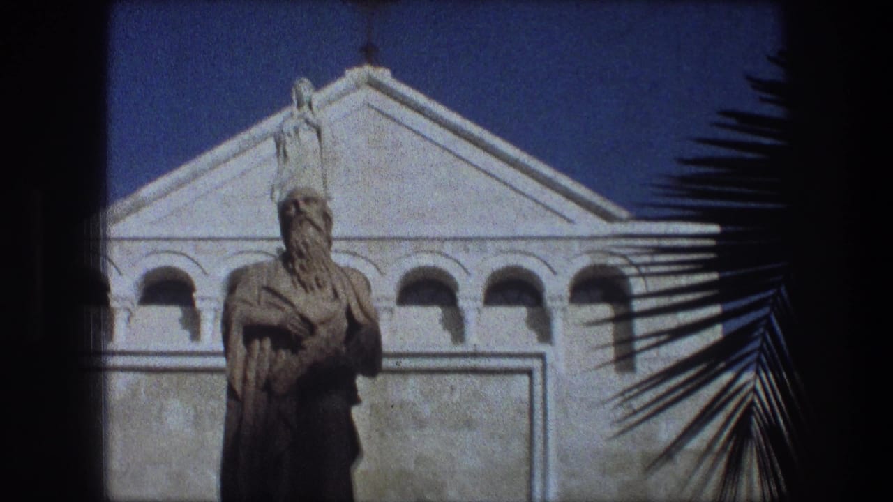 Bearded statue before a pale building facade with arched openings and a small cross.