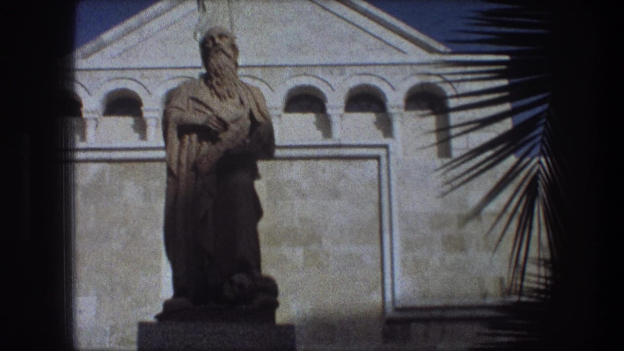 Statue on a pedestal framed by a white facade, blue sky, and palm fronds.