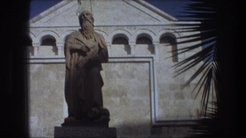 1976 Bethlehem travel frame with a statue before a pale arched facade, palm fronds entering from the right.
