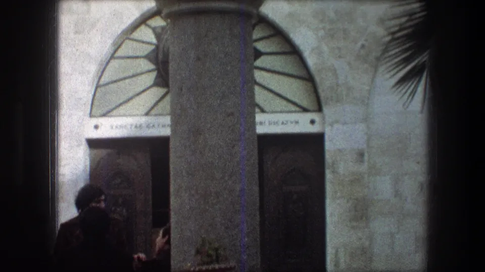 1976 8mm frame from Bethlehem showing robed men near a carved wooden doorway in a pale stone monastery facade.
