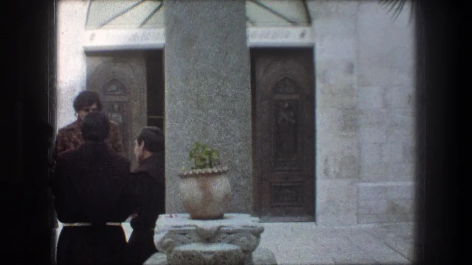 1976 Bethlehem monastery exterior with robed men gathered beside a stone column, carved doors, and a large potted plant.