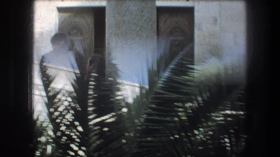 Soft 8mm Bethlehem frame with a robed figure near a stone doorway, partly hidden by palm fronds in the foreground.