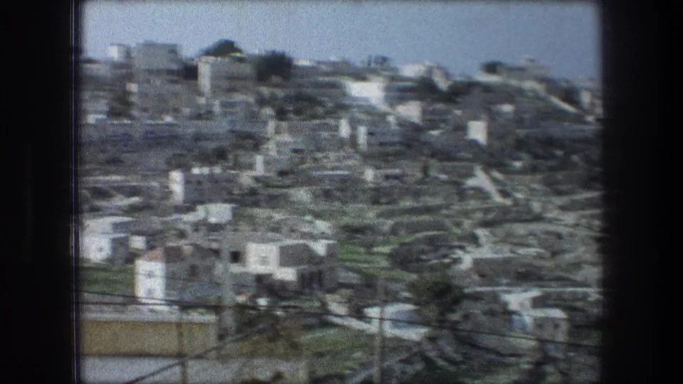 Soft-focus 8mm pan frame of the Bethlehem hillside townscape, with pale buildings, utility lines, and terraced ground.
