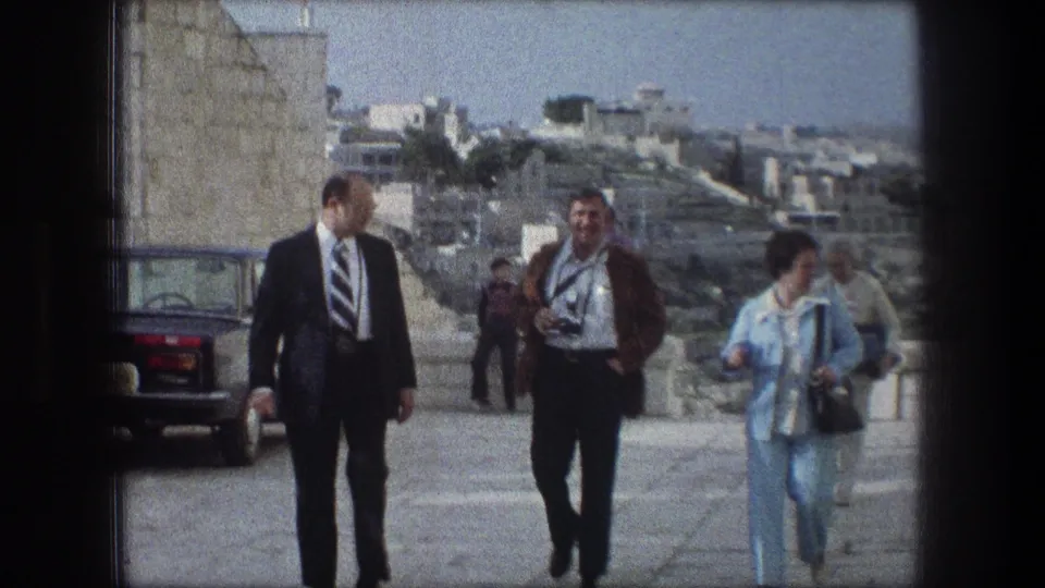 Closer 8mm view of adults walking on a Bethlehem stone path, including a suited man, a man in a brown jacket, and a woman in light blue.