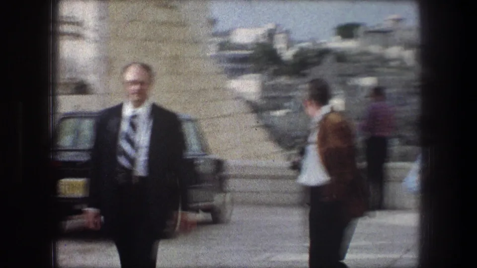 Soft 8mm frame from 1976 Bethlehem with a suited man in the foreground, other adults on the path, and parked cars near hillside buildings.
