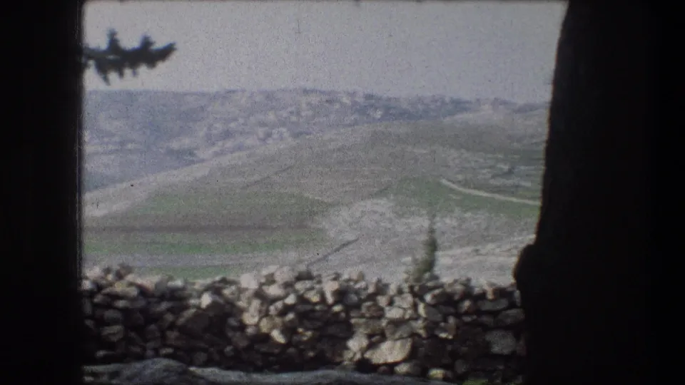 1976 8mm frame of Bethlehem-area hills with a low stone wall in the foreground and distant clustered buildings on the ridge.