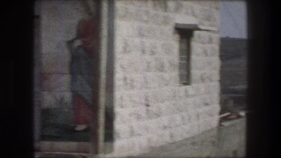 Bethlehem home-movie frame with a painted figure on a stone wall, a small window, and part of a roofline beyond.