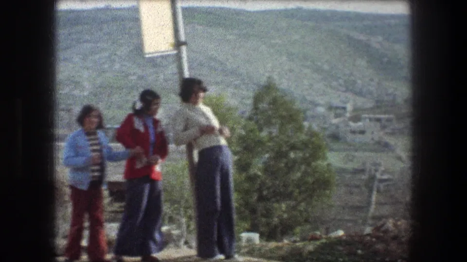 1976 Bethlehem home-movie frame showing three young people near a signpost, with a tree, low buildings, and hills behind them.