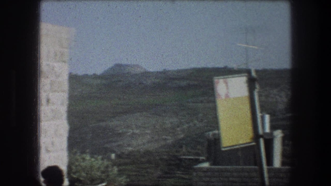 Hillside view through a window with stone wall, yellow sign, rooftops, and distant green slopes.