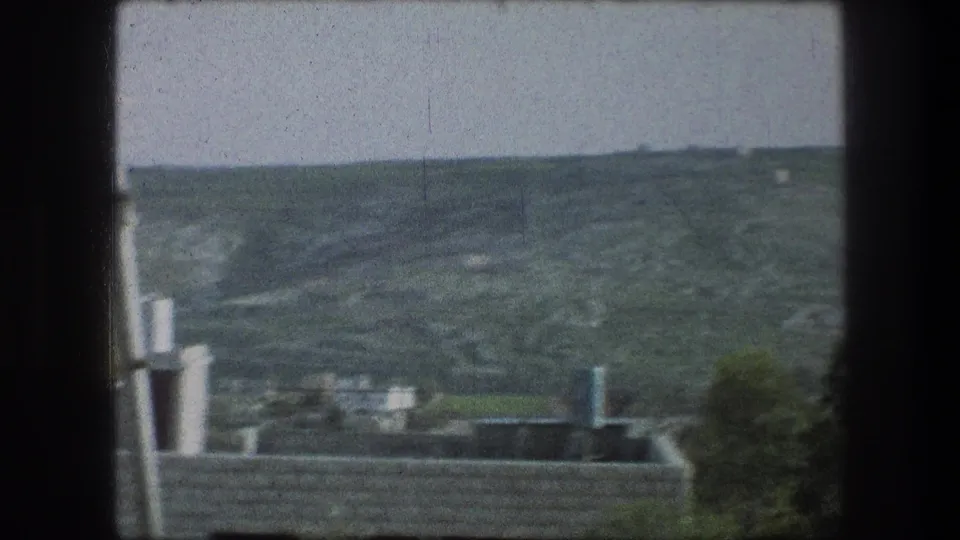 1976 8mm view over Bethlehem rooftops and vent pipes toward scattered hillside houses under a pale sky.