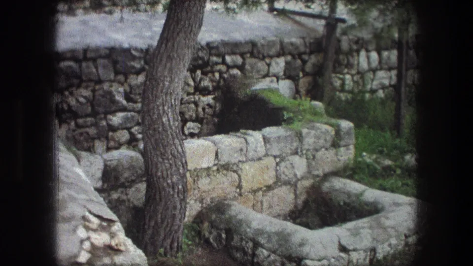 Outdoor Bethlehem scene with a tree, low stone wall, grass, and a rectangular weathered well captured on grainy 8mm film.