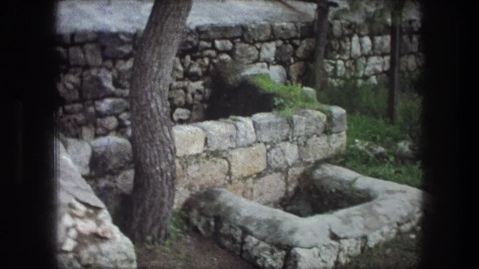 1976 8mm Bethlehem footage showing a weathered stone basin or well near limestone walls, tree trunks, and low greenery.