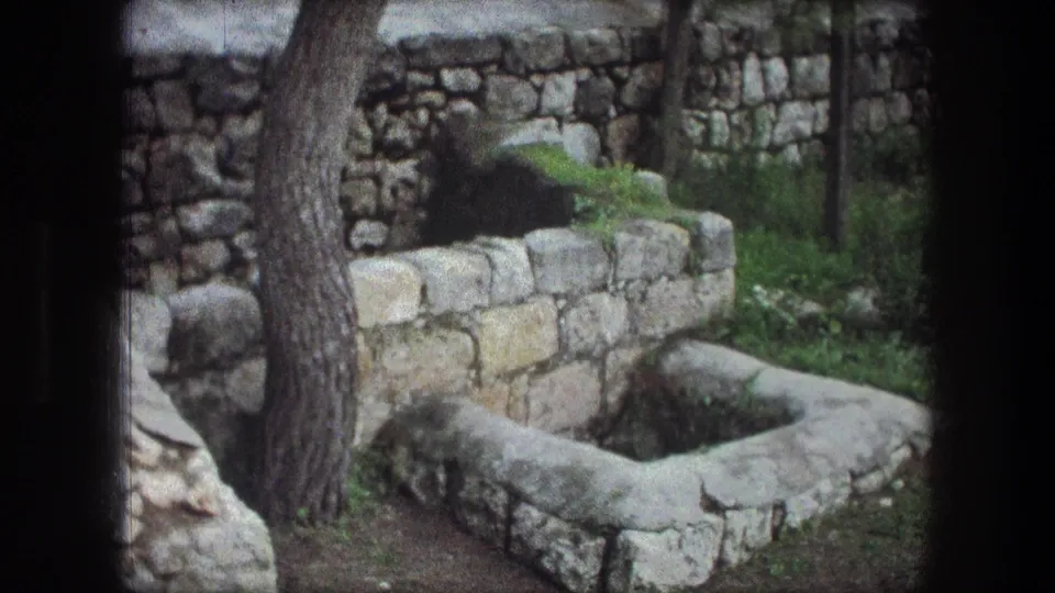 Bethlehem 1976 home-movie frame showing a rough stone basin or well beside a tree and stacked stone wall.