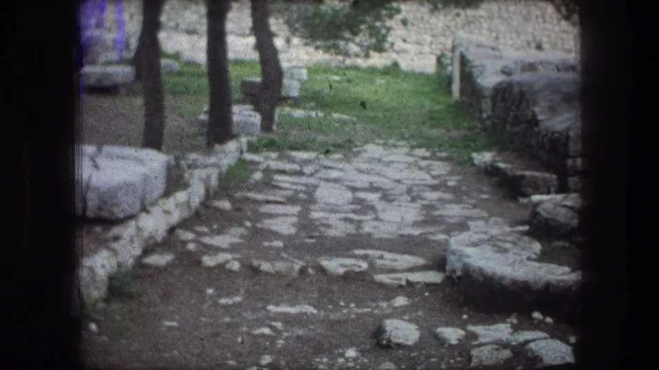1976 8mm Bethlehem frame showing a cobblestone walkway among trees, grass, low stones, and larger weathered stone pieces.