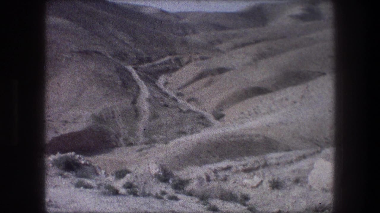 1976 home-movie frame near Bethlehem with a winding mountain road, dry slopes, and a rocky foreground.