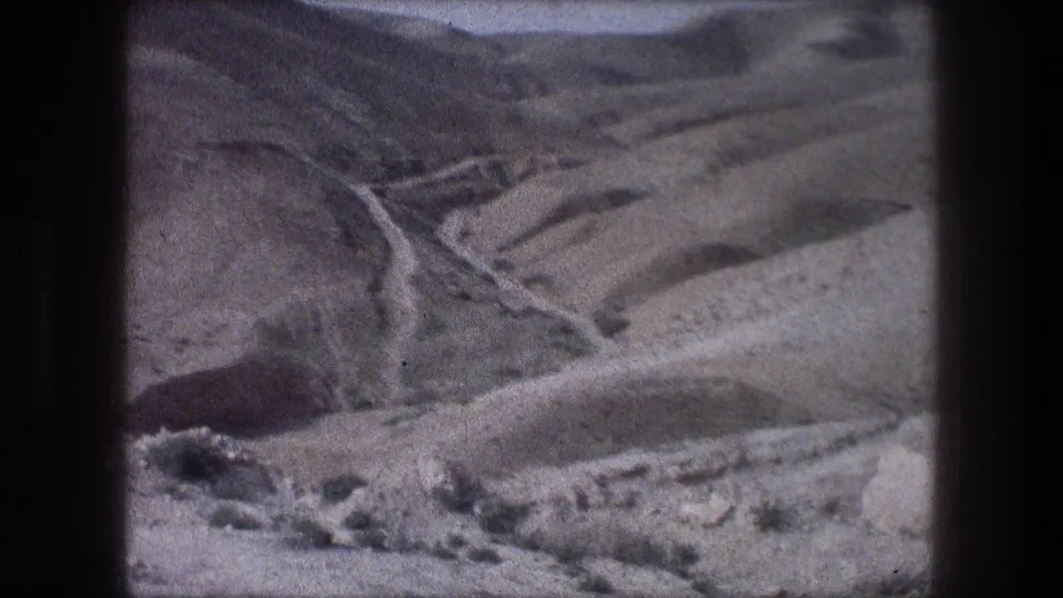 1976 home-movie frame near Bethlehem with a winding mountain road, dry slopes, and a rocky foreground.