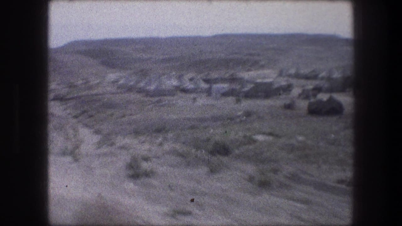 Arid valley with distant tents or low shelters seen through dark film frame edges.