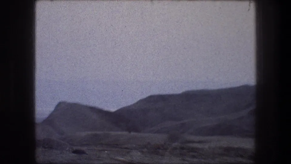 1976 home-movie frame showing low dark mountains and open dry terrain, with subdued color and visible 8mm texture.