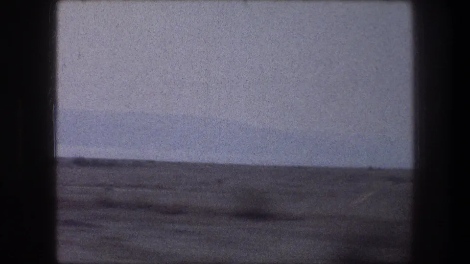1976 Bethlehem-area vehicle-window view of empty arid ground beneath a pale sky, with visible 8mm grain and gate edges.