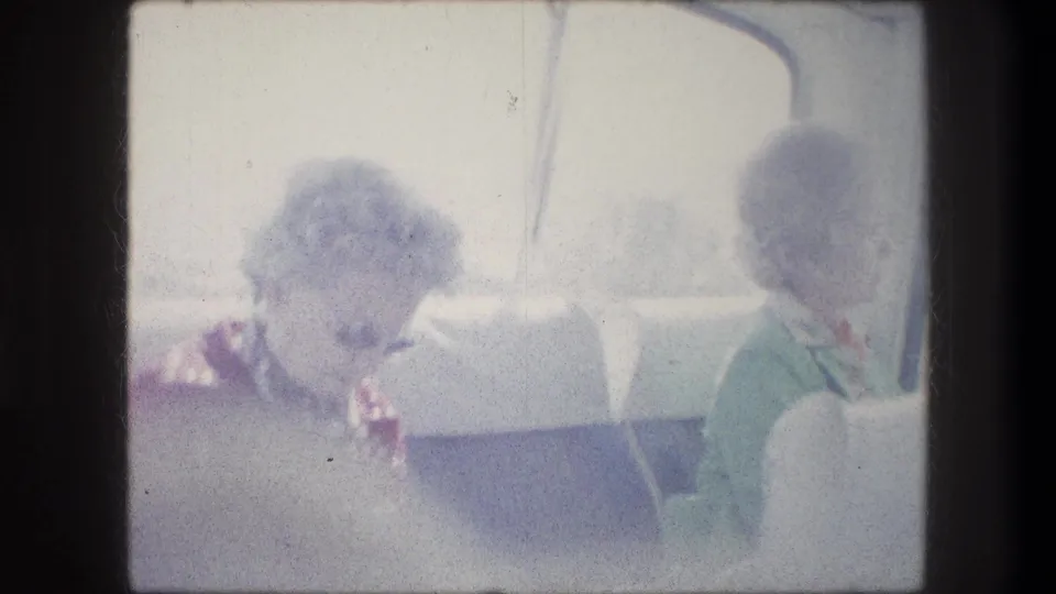 Inside the tour bus, one woman in a patterned top looks down while another in green sits beside the window in faded 1970s 8mm footage.