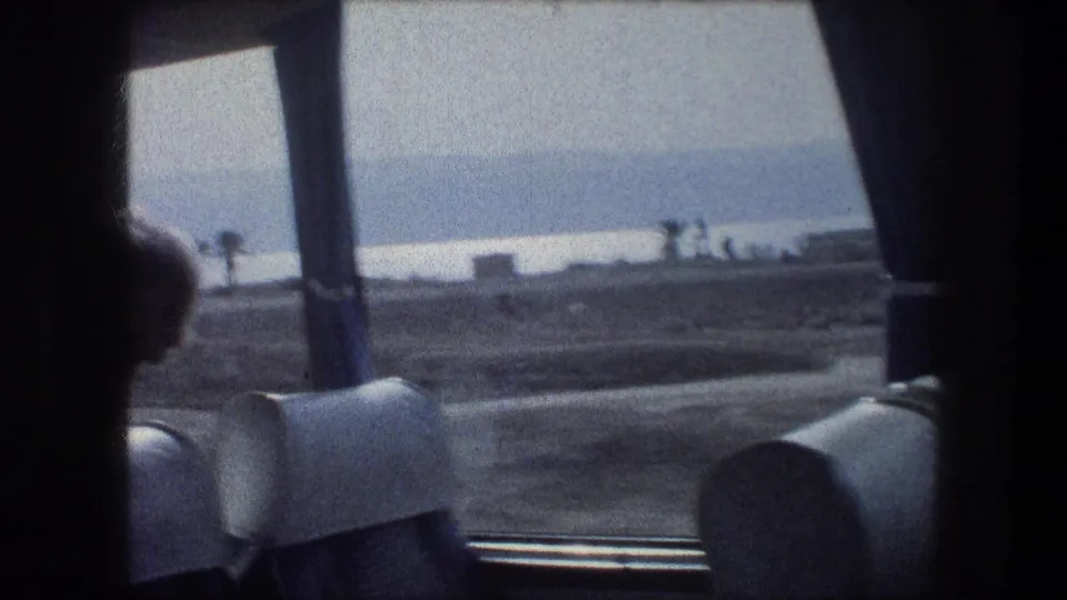 A bus-window view shows seat backs, a dark window frame, hazy open ground, distant water, and small figures outside in the Bethlehem travel footage.