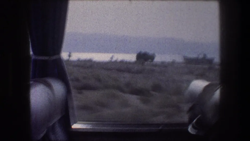 A 1976 bus-window view near Bethlehem, Israel, shows sparse shrubs, low hills, and hazy light beyond the interior curtain.