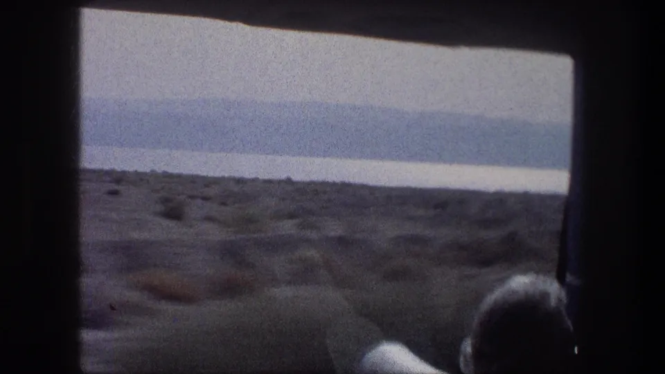 1976 Bethlehem-area passenger POV with the back of a person's head, a seat edge, blue curtain, dry roadside, and distant hills outside.