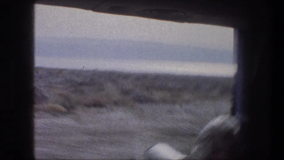 1976 home-movie frame from a moving vehicle, showing sandy coastal terrain and a pale sea horizon through dark window framing.