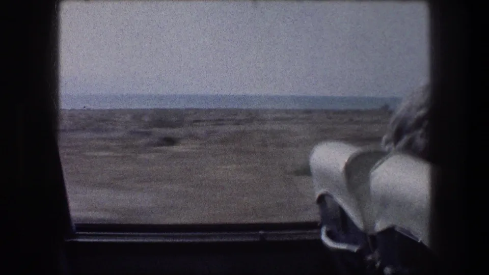 Window-framed 1976 transit view in Israel, with arid coastal ground, distant water, and a white vehicle seat in the foreground.