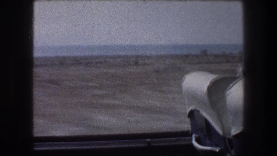 Soft 8mm frame of a 1976 Israel transit-window view, showing scrubby coastal terrain, distant water, and a white seat back.