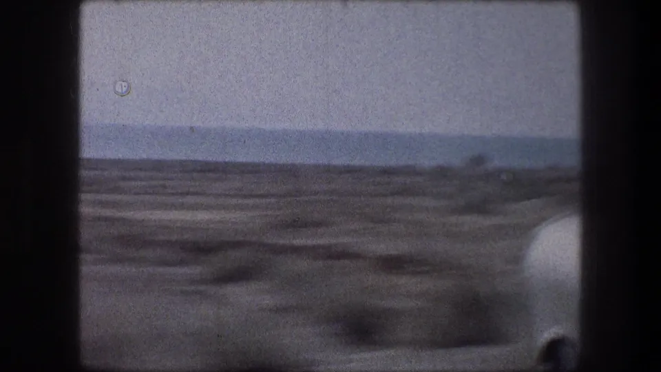 End frame of a 1976 home-movie transit view, with sandy ground, distant water, and dark vehicle framing around the window.