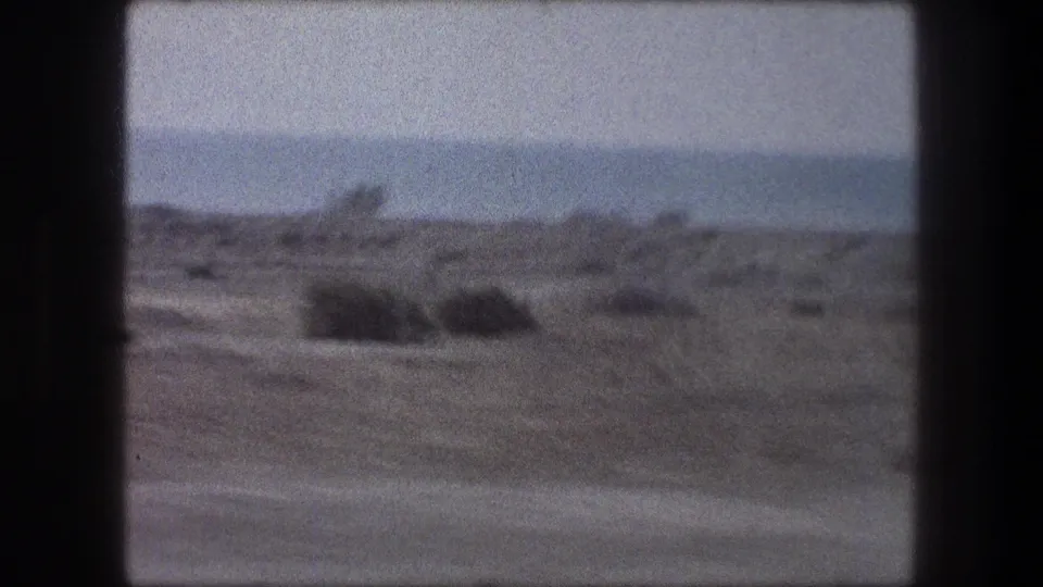Passenger-window view of arid ground, scattered brush, and distant hills in 1976 8mm travel footage.