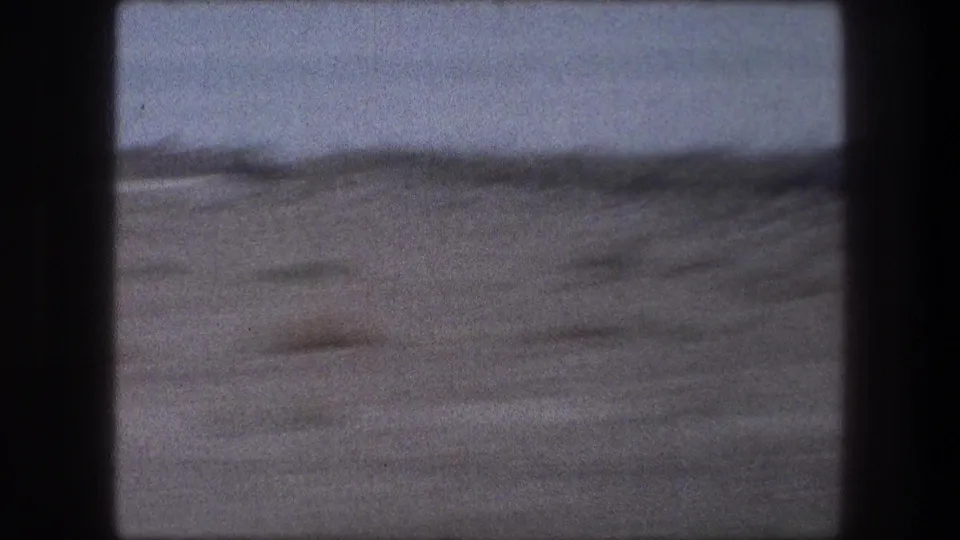 Fast 8mm passenger view of pale sandy ground and desert brush, softened by motion blur and window framing.