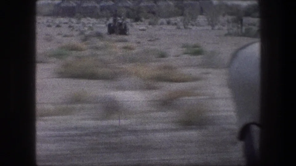 1976 8mm frame of scrubby desert terrain from a moving vehicle, with a partial vehicle edge visible at right.