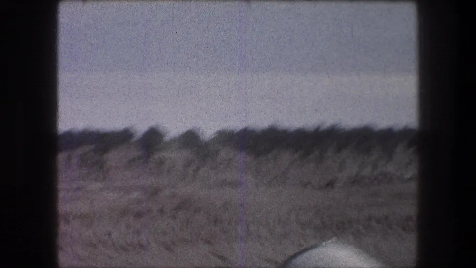 1976 Bethlehem-area home-movie frame showing a dry field, distant shrubs, pale sky, and dark film gate edges.