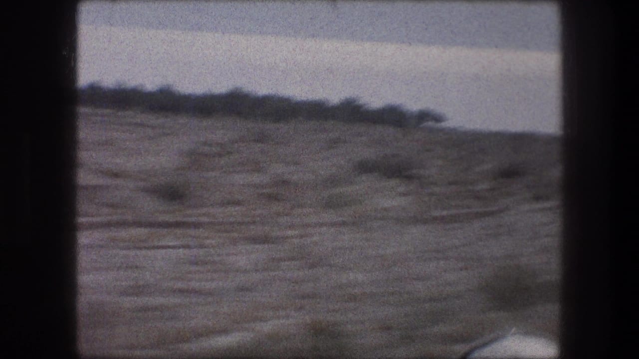 1976 car-window footage near Bethlehem, with dry roadside terrain, sparse shrubs, and a distant low ridge under a pale sky.
