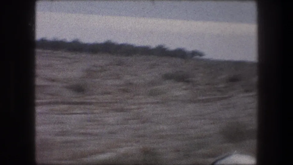 1976 car-window footage near Bethlehem, with dry roadside terrain, sparse shrubs, and a distant low ridge under a pale sky.