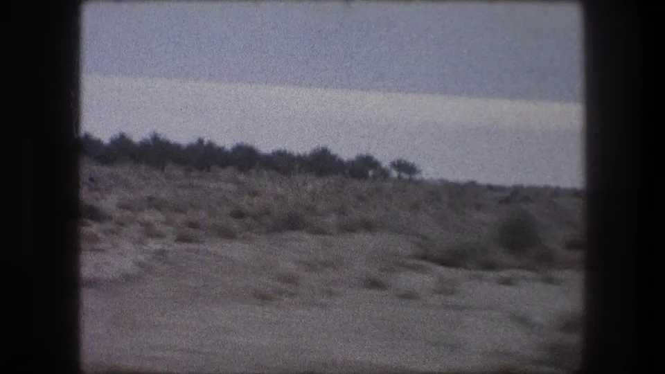 1976 8mm roadside view around Bethlehem showing sandy ground, low brush, and a dark line of distant vegetation.